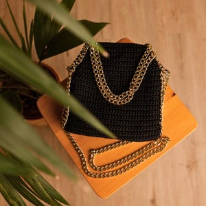 May include: A black, crocheted handbag with a gold chain strap. The bag is square-shaped with a textured surface. The gold chain is draped over the top and extends down the sides, creating a stylish and elegant look. The bag is sitting on a wooden surface.