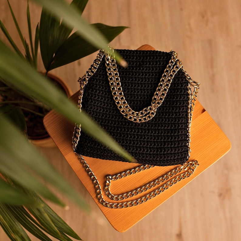 May include: A black, crocheted handbag with a gold chain strap. The bag is square-shaped with a textured surface. The gold chain is draped over the top and extends down the sides, creating a stylish and elegant look. The bag is sitting on a wooden surface.