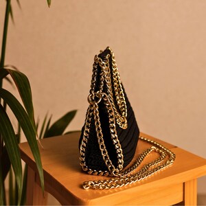 May include: A black, crocheted handbag with a gold chain strap and accents. The bag is sitting on a wooden surface. The gold chain is draped over the bag and extends to the side.