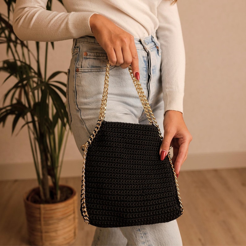 May include: A black crocheted handbag with a gold chain strap. The bag is held by a person wearing a white sweater and blue jeans. The bag has a rectangular shape with rounded corners.