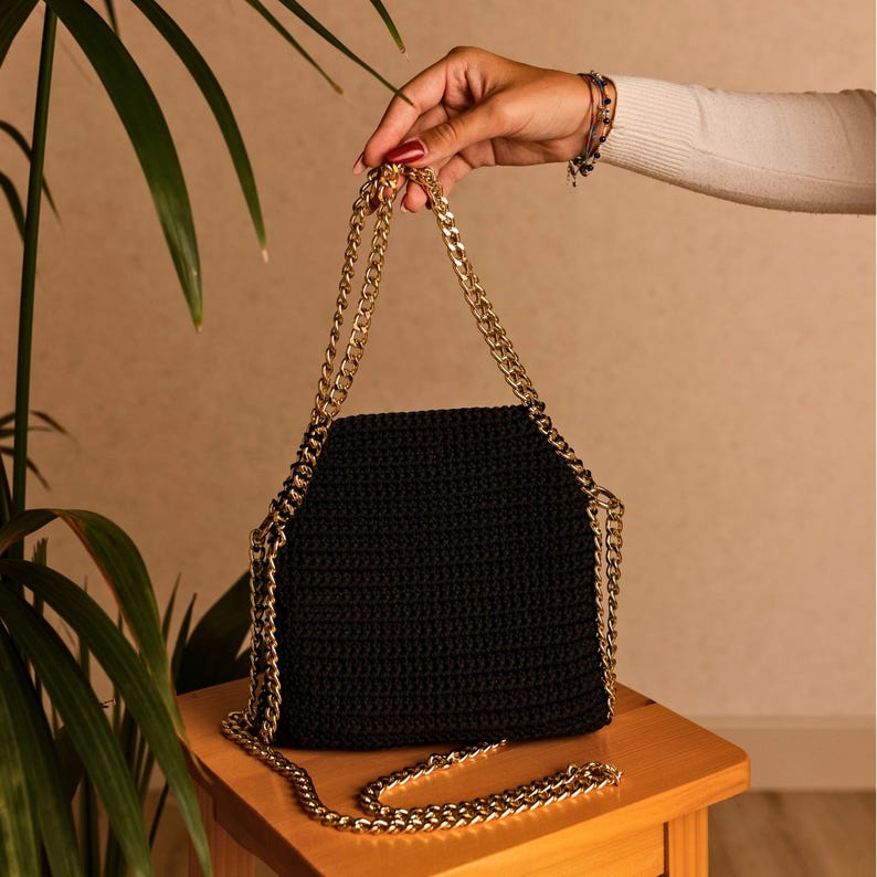 May include: A black crochet handbag with a gold chain strap. The bag is held up by a person's hand. The bag is sitting on a wooden stool. The chain is a gold color.