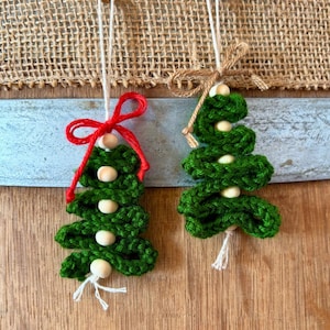 Può includere: Due ornamenti fatti a mano a forma di albero di Natale all'uncinetto verde. Ogni albero è adornato con perline di legno e uno spago per appenderlo. Uno ha un fiocco rosso, l'altro un fiocco di spago naturale. Lo sfondo include una trama di iuta e una superficie in legno.