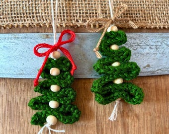 May include: Two handmade green crochet Christmas tree ornaments. Each tree is adorned with wooden beads and a string for hanging. One has a red bow, the other a natural twine bow. The background includes a burlap texture and a wooden surface.