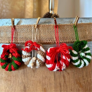Könnte beinhalten: Vier gehäkelte Weihnachtskranz-Ornamente. Jeder Kranz hat eine andere Farbkombination: rot und grün, weiß und braun, rot und weiß und grün und weiß. Jeder Kranz hat eine rote, weiße oder grüne Schnur zum Aufhängen und ist mit einer Schleife und kleinen Glöckchen verziert.