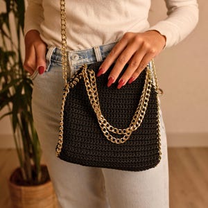 May include: Black crochet crossbody bag with a gold chain strap and accents. The bag is held by a person wearing a white long-sleeve shirt and blue jeans. The bag has a rectangular shape.