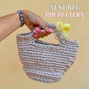 Può includere: Una borsa a nido beige, lavorata a mano all'uncinetto, con forma arrotondata e manici robusti. La borsa è adornata da un fiore giallo e il testo "NEST BAG PDF PATTERN" è visualizzato in alto a destra.