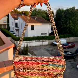 Può includere: Una borsa rettangolare all'uncinetto multicolore con una tracolla a catena dorata. La borsa presenta un vivace mix di fili rossi, rosa, gialli e blu. La catena dorata è attaccata agli angoli superiori della borsa.