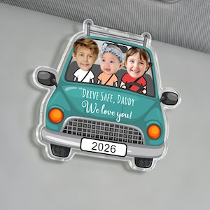Pode incluir: Um acessório de viseira solar em forma de carro turquesa com os rostos de três crianças no interior. O texto "DRIVE SAFE, DADDY We love you!" está impresso no carro. O ano "2026" é exibido.
