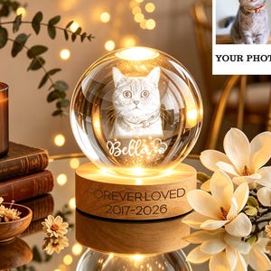 Puede incluir: Una esfera de cristal transparente con un retrato de gato y el nombre "Bella" grabado. La esfera está sobre una base de madera con la inscripción "FOREVER LOVED 2017-2026". Una vela encendida, libros y flores en el fondo.