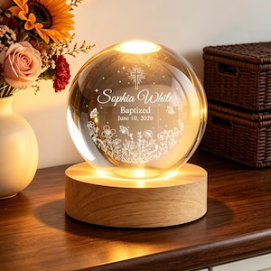 May include: A clear glass sphere lamp on a round wooden base, illuminated from within. The sphere features an etched cross, the name Sophia White, the word Baptized, the date 10 June 2026, and a floral design with butterflies.