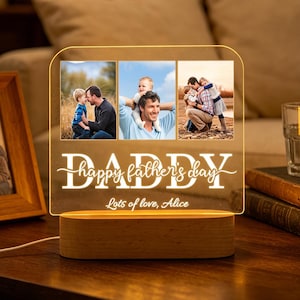 Può includere: Un regalo personalizzato per la festa del papà con tre foto di famiglia, illuminato da una calda luce. Il pannello in acrilico mostra le parole "DADDY happy father's day" e "Lots of love, Alice" su una base di legno.