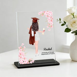 Puede incluir: Placa de graduación de acrílico transparente con una graduada con birrete y toga, sosteniendo un diploma. La placa está decorada con detalles florales rosas y el texto "Rachel, University of Chicago, Department of Finance."