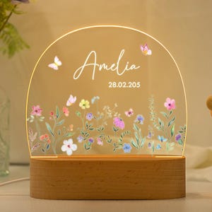 May include: A white acrylic night light with a floral design and the name Amelia and the date 28.02.205. The light is on a wooden base.