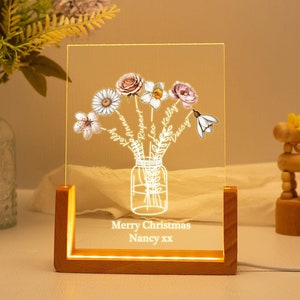 May include: A personalized LED night light with a wooden base. The clear acrylic panel features a floral bouquet design with names and the text "Merry Christmas Nancy xx". The flowers include roses, daisies, and other blooms.