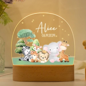 Pode incluir: Uma luz noturna de madeira com uma cúpula de acrílico transparente que apresenta uma cena de selva caprichosa com uma zebra, uma girafa, um elefante, uma raposa e um leão. A cúpula é iluminada com luzes LED branco quente e a base é gravada com o nome "Alice" e a data "06.14.2024".