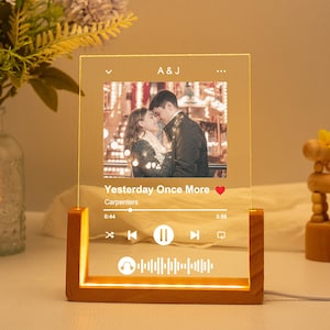 Könnte beinhalten: Ein Holzrahmen mit einer Glasfront, der ein Foto eines Paares und eine Musikplayer-Oberfläche zeigt. Der Musikplayer zeigt den Song "Yesterday Once More" von The Carpenters, mit einem roten Herz-Symbol und einer Fortschrittsanzeige. Der Rahmen hat eine helle Holzbasis und einen hellbraunen Hintergrund.