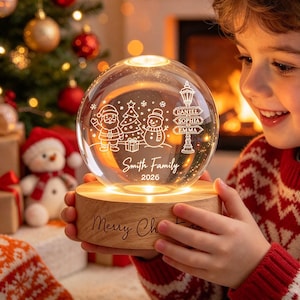 Könnte beinhalten: Ein Nachtlicht in Form einer klaren Glaskugel auf einem runden Holzsockel. Das Glas zeigt ein eingraviertes Design mit Weihnachtsmann, Tannenbaum, Schneemann, einem Straßenschild mit den Namen Daniel, Sophia, Emma und Smith Family 2026.