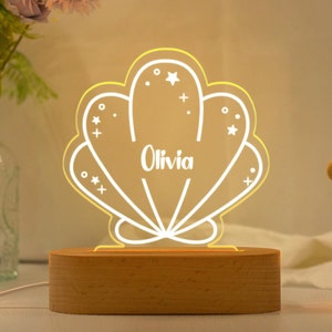 Puede incluir: Una luz nocturna de madera con forma de concha con un contorno blanco y el nombre "Olivia" en el interior. La concha tiene un brillo amarillo y está rodeada de pequeñas estrellas blancas.