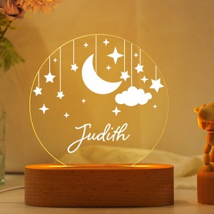 May include: A white acrylic night light with a moon, stars, and clouds design. The light has a wooden base and is illuminated with a warm yellow glow. The name "Judith" is engraved on the light.