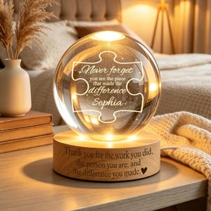 May include: A lit crystal ball night light on a wooden base. The ball features a puzzle piece design with the words "Never forget you are the piece that made the difference Sophia." The base reads "Thank you for the work you did, the person you are, and the difference you made."