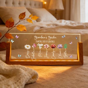 May include: A personalized LED light with a wooden base. The clear acrylic panel displays the text "Grandma's Garden" and "Planting Seeds of Greatness", along with names and floral illustrations. Butterflies are scattered across the panel.