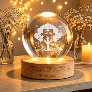 Può includere: Una sfera di vetro trasparente con un disegno ad albero fatto di pezzi di puzzle, illuminata dall'interno. La sfera poggia su una base di legno con la scritta "Thank you for being such an important piece of my story" e il nome "Amelia".