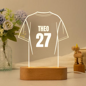 May include: A white outline of a jersey with the name "THEO" and the number "27" on the back. The jersey is illuminated from behind and sits on a wooden base.