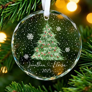 May include: A clear, round Christmas bauble featuring a painted green Christmas tree, red baubles, and falling snow. The names "Jonathan & Elvira" and the year "2025" are written on the bauble. A white ribbon is attached for hanging.