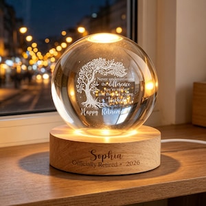 Op de afbeelding: Een heldere glazen bolvormige lamp op een ronde, lichtgekleurde houten voet. De bol bevat een gegraveerde boom en de tekst: Never underestimate the difference you've made and the lives you've touched. Happy Retirement. Op de voet staat: Sophia Officially Retired 2026.