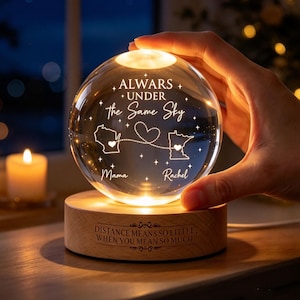 Puede incluir: Una luz nocturna de esfera de cristal transparente sobre una base redonda de madera. La esfera está grabada con el texto "ALWAYS UNDER the Same Sky", dos contornos de estados conectados por una línea punteada con un corazón, y los nombres "Mama" y "Rachel". La base presenta el texto grabado "DISTANCE MEANS SO LITTLE, WHEN YOU MEAN SO MUCH."