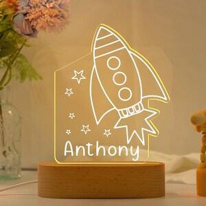 Peut inclure: Une fusée blanche avec des étoiles et le nom Anthony sur une base en bois. La fusée est dessinée en blanc et a une lueur jaune.