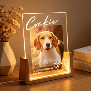 Può includere: Una cornice portafoto rettangolare in acrilico con base in legno illuminata. La cornice mostra la fotografia di un cane beagle con il nome Cookie stampato in alto e il testo IN LOVING MEMORY 2001-2025 con una zampa grafica in basso.