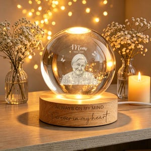Peut inclure: Une sphère de verre transparente avec un portrait de femme, le mot "Mom" et des dates, repose sur une base en bois. La base est gravée de "ALWAYS ON MY MIND" et "Forever in my heart". La sphère est éclairée de l'intérieur, créant une lueur chaleureuse.