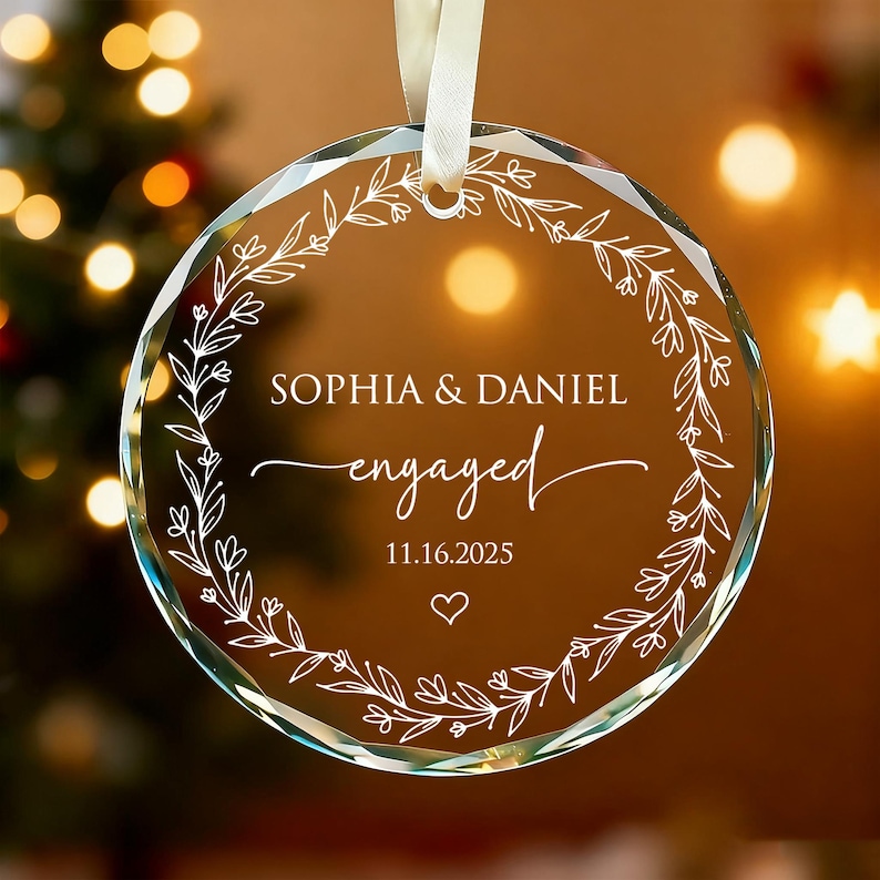 May include: A clear, round glass ornament with a white ribbon for hanging. The ornament features the names "SOPHIA & DANIEL" and the word "engaged" in script, along with the date "11.16.2025" and a heart symbol, surrounded by a floral wreath.