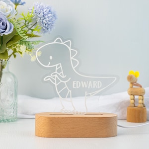 May include: A clear acrylic night light featuring a white line-art illustration of a dinosaur with the name EDWARD engraved on its body, mounted on a smooth, oval-shaped light-coloured wooden base with a power cord extending from the side.