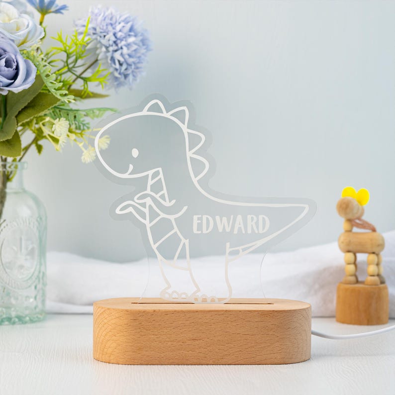 May include: A clear acrylic night light featuring a white line-art illustration of a dinosaur with the name EDWARD engraved on its body, mounted on a smooth, oval-shaped light-coloured wooden base with a power cord extending from the side.