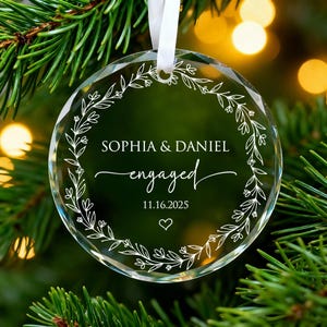 May include: A clear, round glass Christmas ornament with the names "SOPHIA & DANIEL" and the word "engaged" in elegant script. The date "11.16.2025" and a heart are also featured. A white floral wreath design surrounds the text.