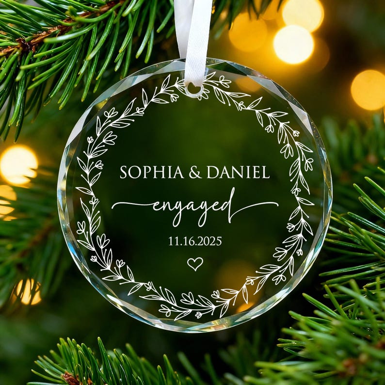 May include: A clear, round glass Christmas ornament with the names "SOPHIA & DANIEL" and the word "engaged" in elegant script. The date "11.16.2025" and a heart are also featured. A white floral wreath design surrounds the text.