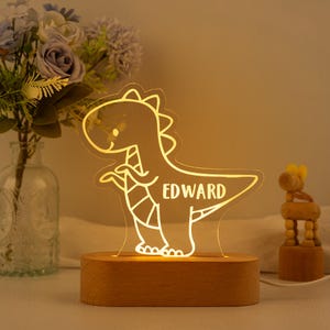 May include: A clear acrylic dinosaur-shaped night light with a warm yellow glow, featuring the name EDWARD etched into the centre. The light is mounted on a smooth, oval-shaped light-coloured wooden base, creating a soft illumination effect.