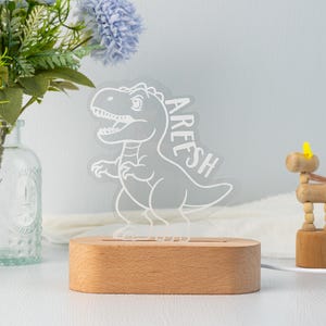 May include: A white dinosaur night light with the name "AREEH" written on it. The light is on a wooden base.