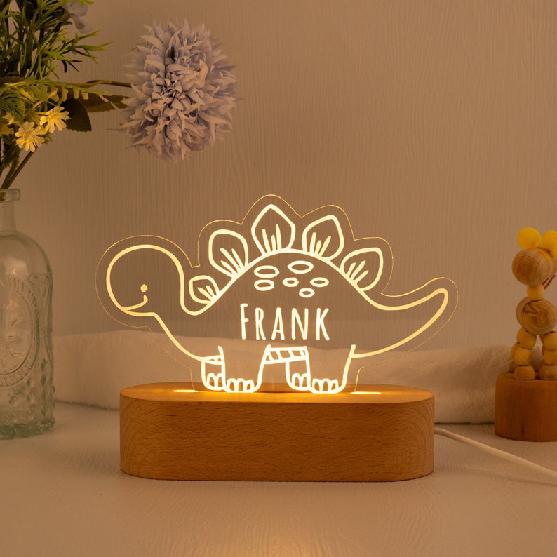 May include: A clear acrylic dinosaur-shaped night light featuring an etched outline of a stegosaurus with the name FRANK engraved in the centre. The acrylic piece is mounted on a smooth, oval-shaped light-coloured wooden base that illuminates.
