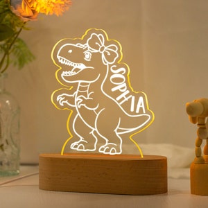 May include: A wooden night light shaped like a dinosaur with a bow on its head. The dinosaur is lit up with a warm yellow glow and the name "Sophia" is written on its side.