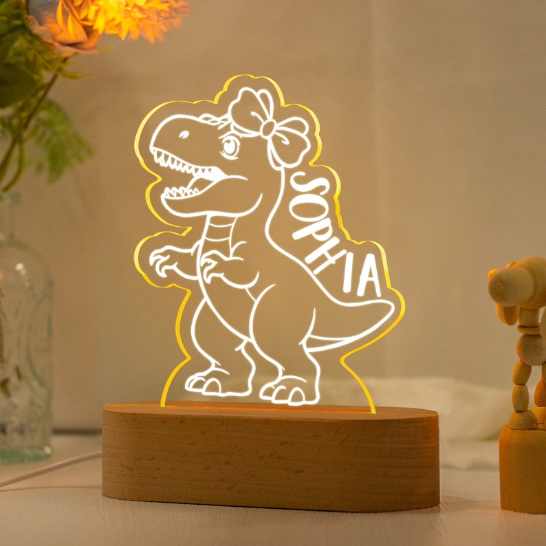 May include: A wooden night light shaped like a dinosaur with a bow on its head. The dinosaur is lit up with a warm yellow glow and the name "Sophia" is written on its side.