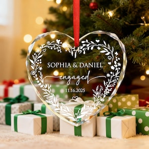 May include: Clear heart-shaped ornament with the names "Sophia & Daniel" and the word "engaged" in script. The ornament has a floral design and the date "11.16.2025". A red ribbon is attached for hanging. The ornament is in front of a Christmas tree and wrapped gifts.