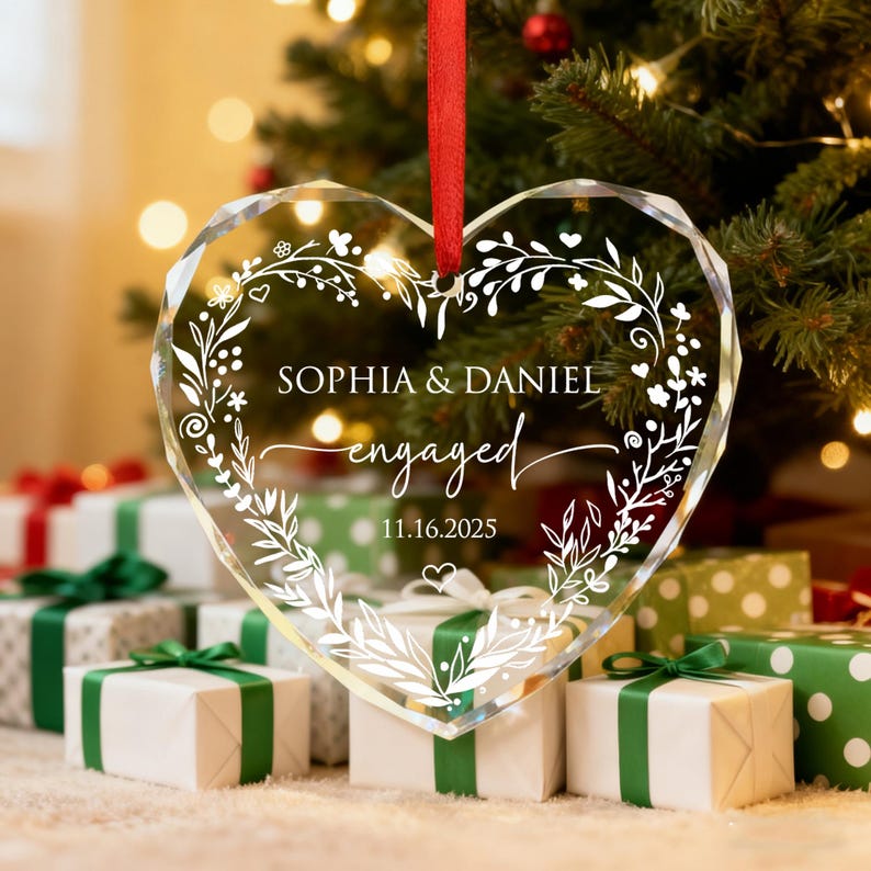 May include: Clear heart-shaped ornament with the names "Sophia & Daniel" and the word "engaged" in script. The ornament has a floral design and the date "11.16.2025". A red ribbon is attached for hanging. The ornament is in front of a Christmas tree and wrapped gifts.