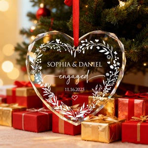 May include: A clear, heart-shaped ornament with the names "SOPHIA & DANIEL" and the word "engaged" in elegant script. The ornament is adorned with a white floral design and the date 11.16.2025. A red ribbon hangs from the top. Presents are in the background.