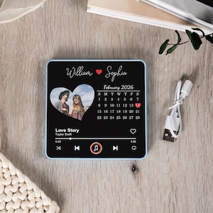 Puede incluir: Un reproductor de m&uacute;sica negro personalizado con un borde azul. Presenta una foto en forma de coraz&oacute;n, los nombres "William" y "Sophia", y un calendario de febrero de 2026. La pantalla muestra una interfaz de reproductor de m&uacute;sica con la canci&oacute;n "Love Story".