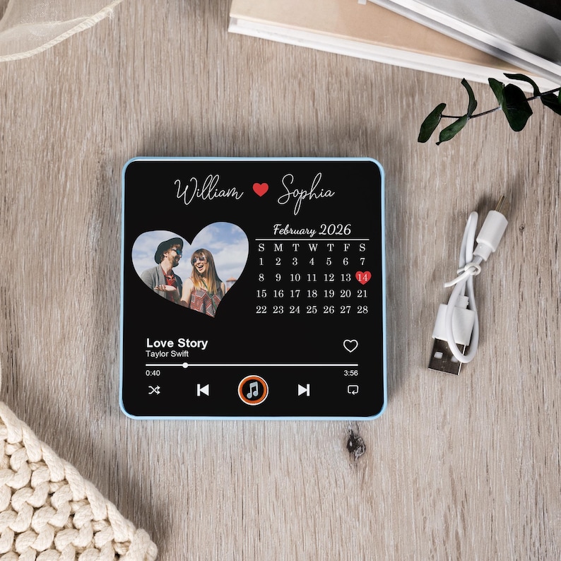 Puede incluir: Un reproductor de m&uacute;sica negro personalizado con un borde azul. Presenta una foto en forma de coraz&oacute;n, los nombres "William" y "Sophia", y un calendario de febrero de 2026. La pantalla muestra una interfaz de reproductor de m&uacute;sica con la canci&oacute;n "Love Story".