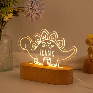 May include: A clear acrylic dinosaur-shaped night light featuring the name FRANK etched into the centre. The dinosaur design is illuminated with a warm glow and is mounted on a smooth, oval-shaped light-coloured wooden base with a power cord.