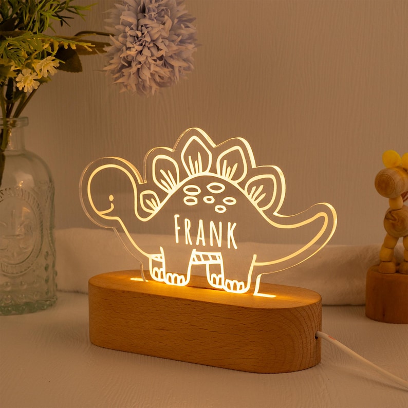 May include: A clear acrylic dinosaur-shaped night light featuring the name FRANK etched into the centre. The dinosaur design is illuminated with a warm glow and is mounted on a smooth, oval-shaped light-coloured wooden base with a power cord.